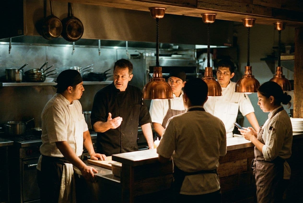 A chef in a black coat talking to cooks and servers gathered at the pass before service.